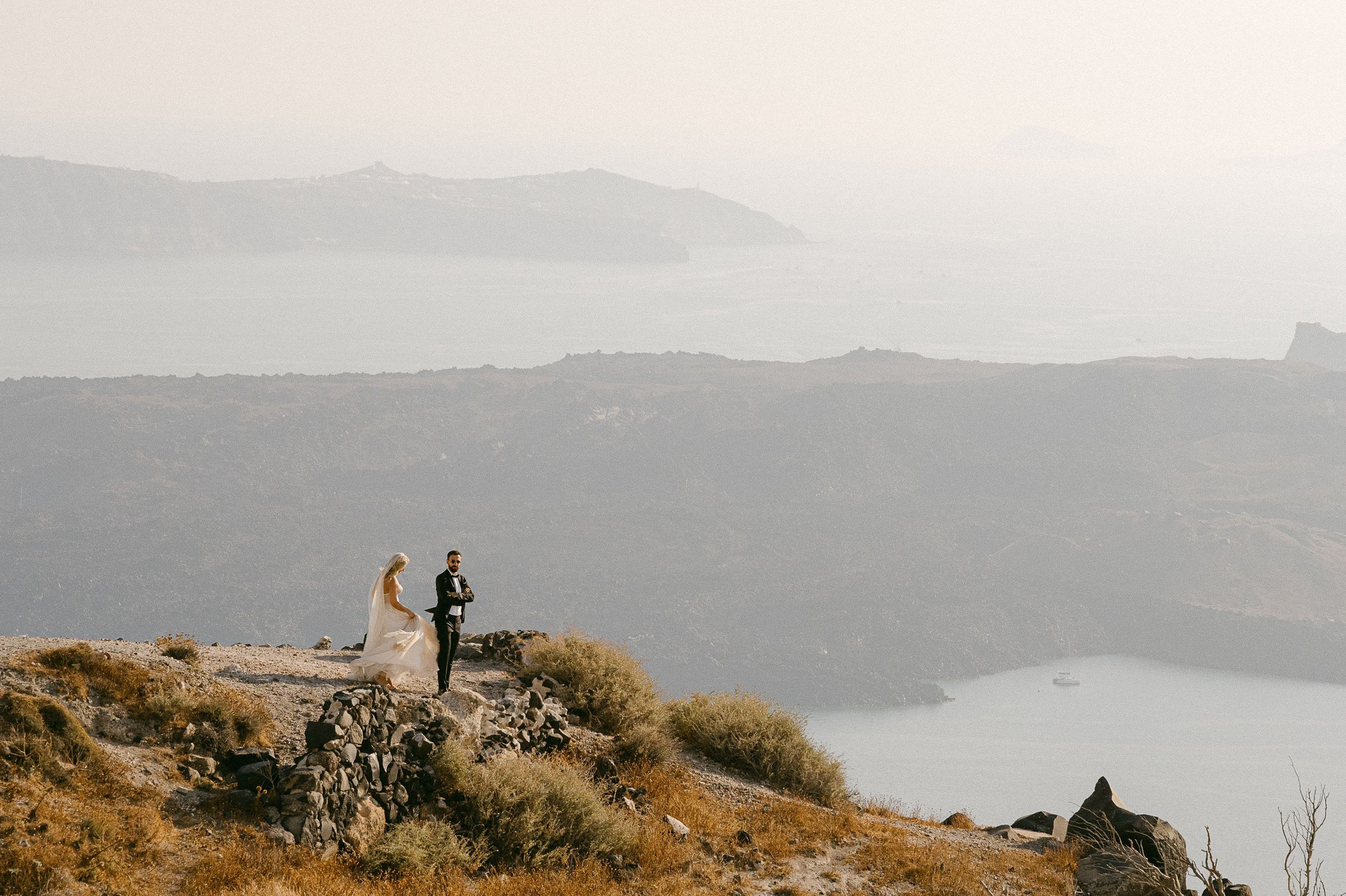 Margi and Nikos standing together on a rocky Santorini ridge above the sea