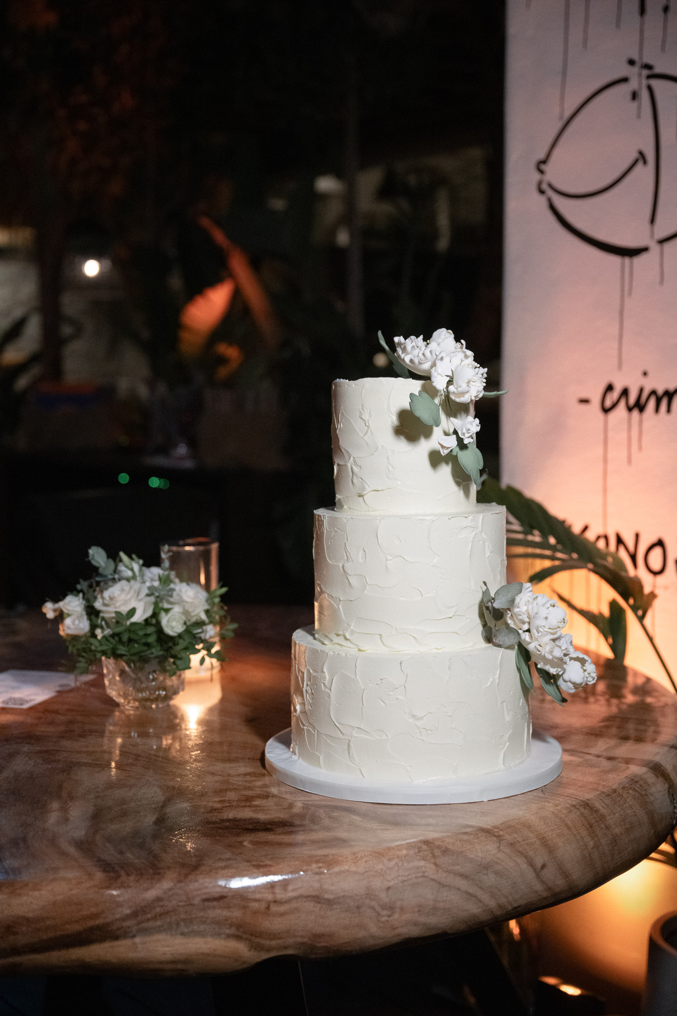White tiered wedding cake decorated with pale flowers at a candlelit Mykonos reception.