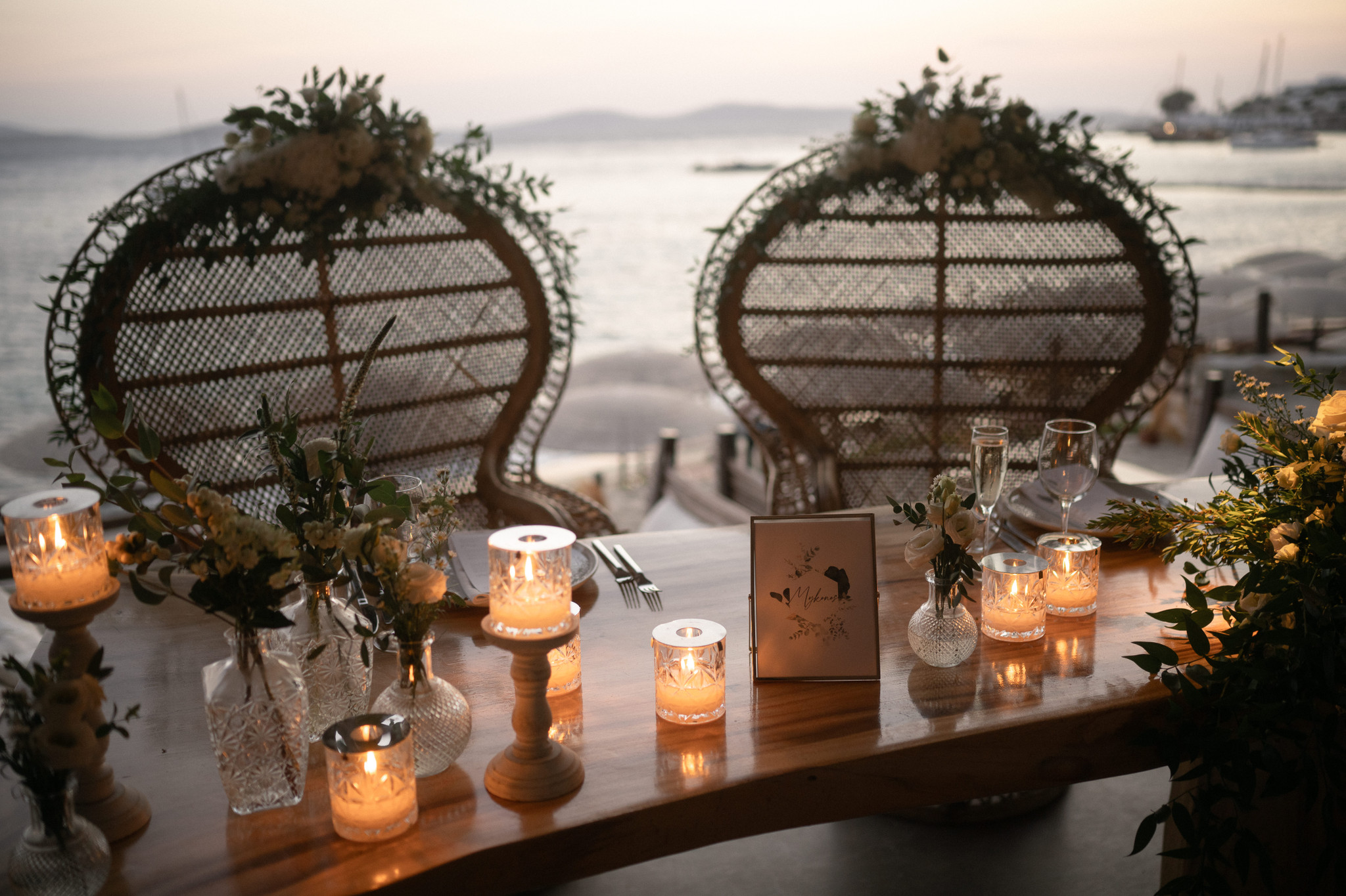 Sunset reception table with candles, floral arrangements and wicker chairs facing the Aegean in Mykonos.