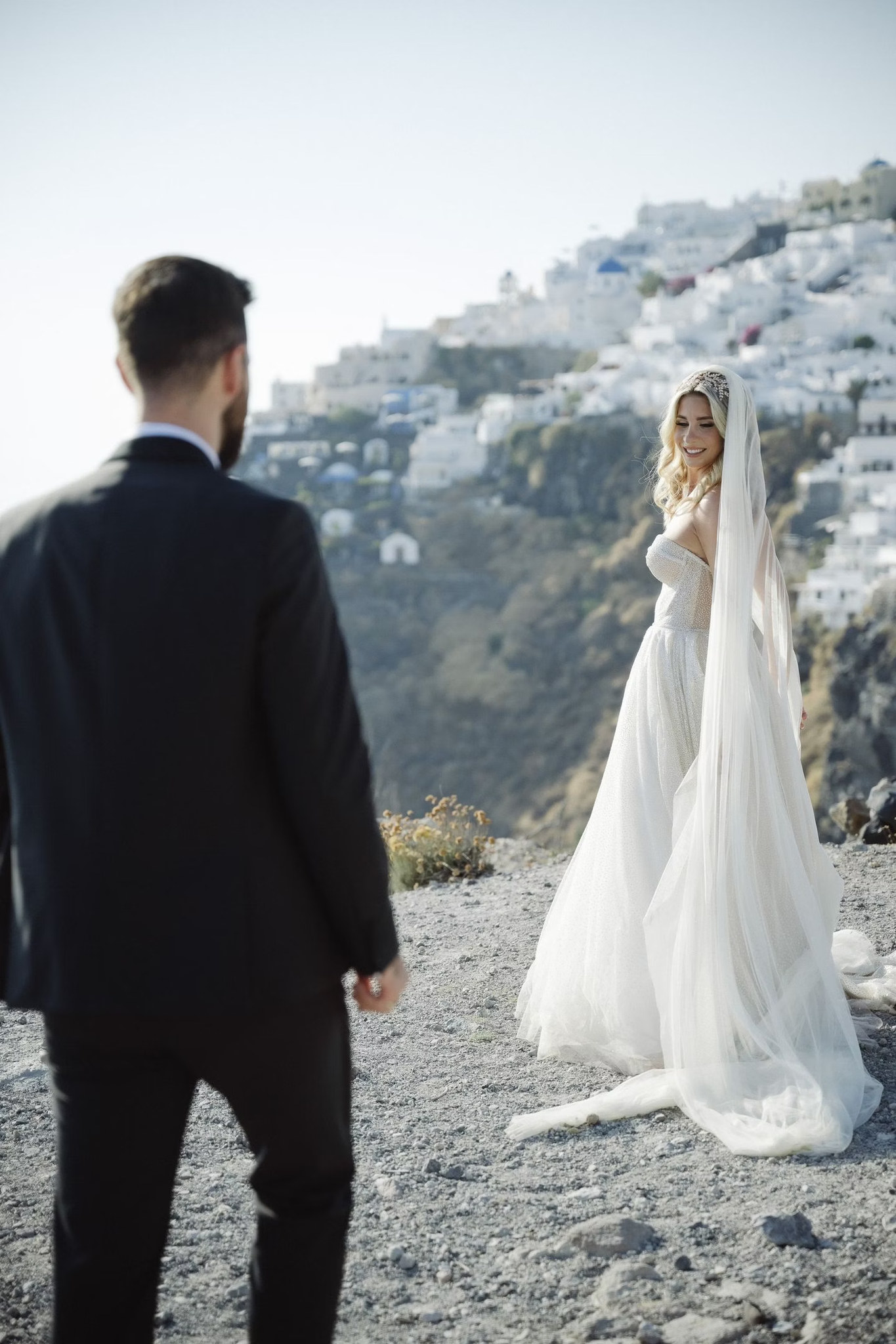 Nikos in the foreground as Margi turns toward him, with Santorini’s whitewashed hillside behind her