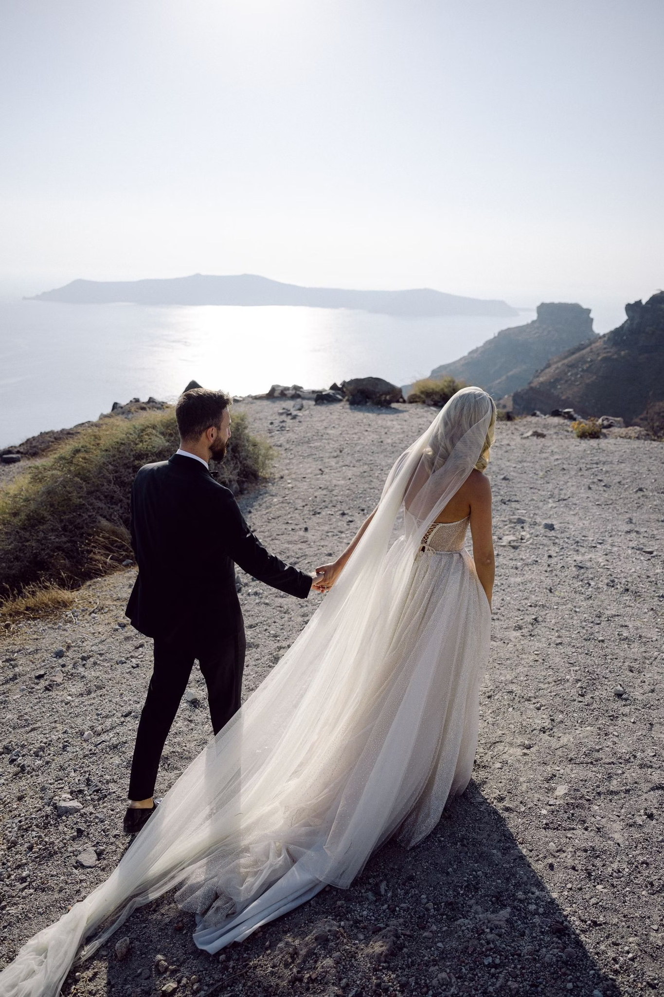 Margi and Nikos walking on a rocky Santorini path, her wedding dress and veil flowing behind her